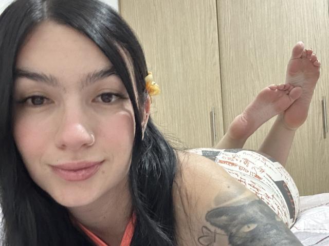 EvaaFarix, avec ses cheveux noirs, son anneau de nez et son bras tatoué, allongée sur un lit, montrant la plante de ses pieds, portant un short à motifs, souriant doucement..