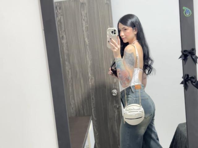 EvaaFarix with long black hair, white tank top, and blue jeans, showing tattooed arm, holding phone, looking back, holding white Chanel purse, standing in front of mirror.