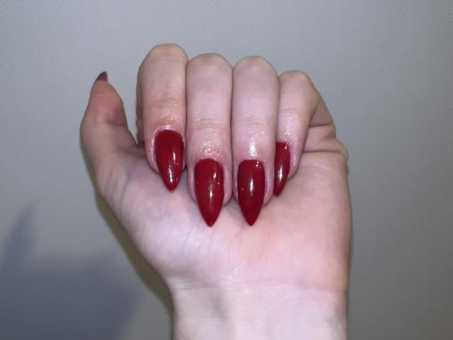 La mano de EvaaFarix, con uñas rojas brillantes y en forma de almendra, dedos ligeramente curvados y un tono de piel claro. Sus uñas son largas y puntiagudas, mostrando un acabado brillante..