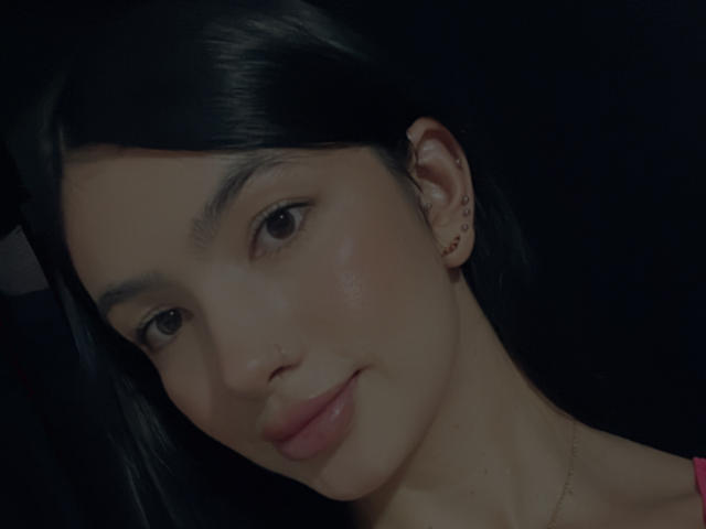 EvaaFarix tiene una piel lisa de color marrón claro, cabello negro y lacio, pequeños aretes y una ligera sonrisa. Sus labios rosados están relajados, y mira suavemente a la cámara..