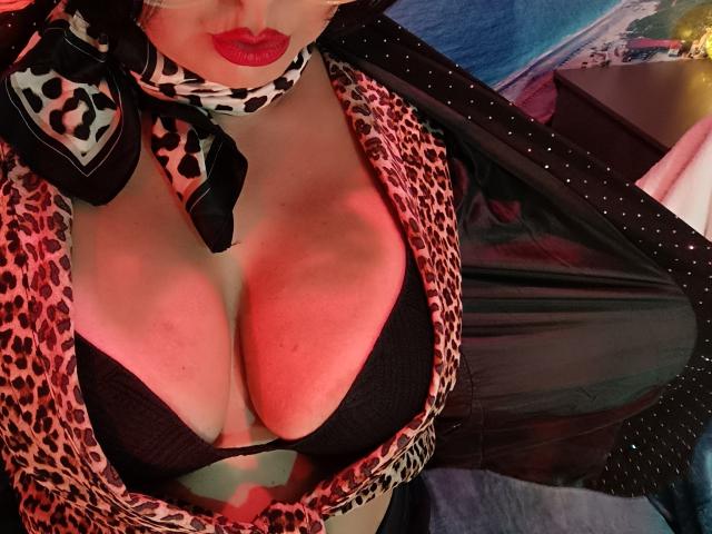 AmberSunshine with big tits, wearing a leopard print top, black bra, and matching scarf, has red lipstick and a black, shiny, polka-dotted jacket open. Boob cleavage visible.