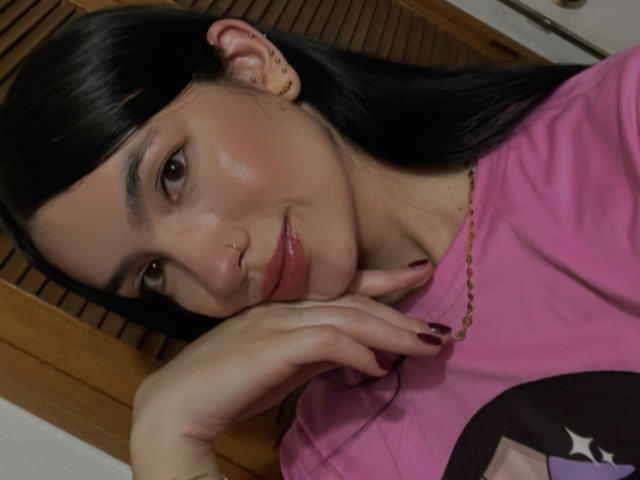 EvaaFarix mit schwarzen Haaren, einem pinken Hemd und einer goldenen Kette, die ihr Kinn in die Hand legt, mit voller Lippe, dunklem Nagellack, braunen Augen und einem leichten Lächeln..