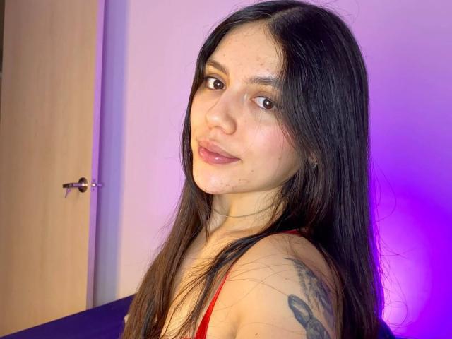 SophiaMarx with long black hair, light skin, subtle makeup, red tank top, small tattoo on right shoulder, looking over shoulder, slight smile, purple and pink lighting.