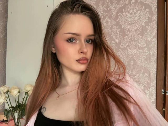 JungSantago with long, straight, reddish-brown hair, pinkish-nude lipstick, subtle blush, black tank top, pink jacket, small tattoo on left chest, gold necklace, white roses in background.