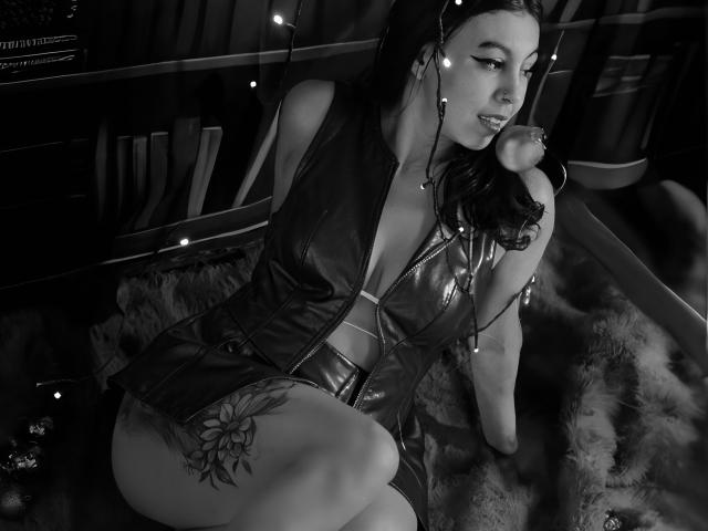NenaBlack mit dunklem Haar, die ein ledernes, mit Reißverschluss versehenes Outfit trägt, sitzt auf einem flauschigen Teppich, hat Weihnachtsbeleuchtung im Haar, ein Tattoo an der Oberschenkel und blickt zur Seite, während sie eine Pfeife hält..