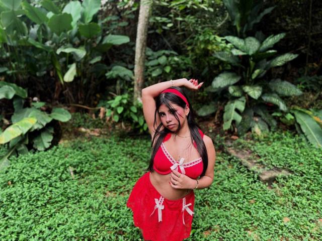 CelesteMorel with medium tits in a red bra and skirt, arm raised, standing on green grass, wearing a red headband, in a lush forest setting.