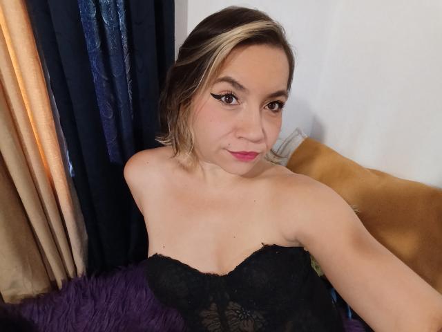 MsJustineBeaux with blonde and brown hair, wearing a black lace strapless top, showing her small tits, pink lipstick, and winged eyeliner, sitting on a purple furry pillow.