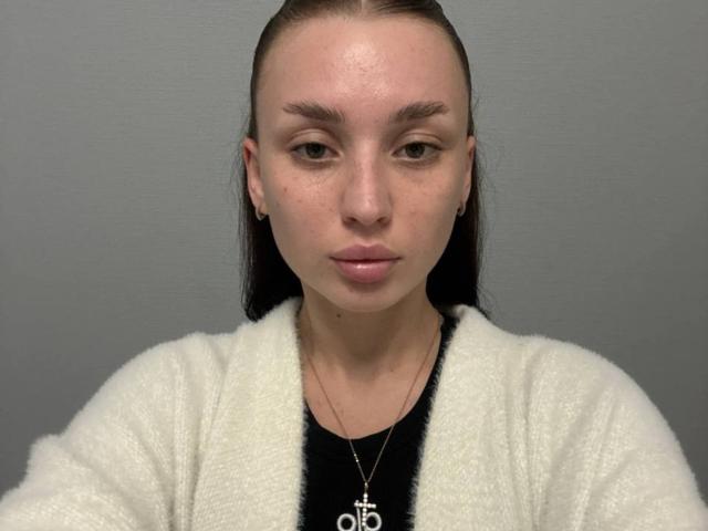 JaynaFeeler ha un viso serio, la pelle chiara, gli occhi marroni e i capelli castani lisci. Indossa un cardigan bianco e morbido sopra una maglietta nera, con una collana d'argento..