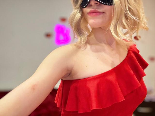 MiyaLiya with blonde wavy hair, wearing a black blindfold and a red ruffled one-shoulder dress, showing her tits and armpit in a selfie.
