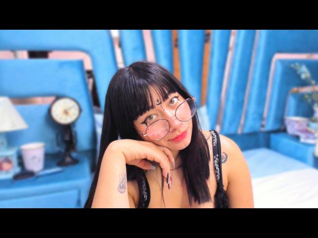 Genesiss with black hair, pink glasses, and a cute face, resting her head on her hand, showing a tattoo on her wrist, wearing a black lace top, in a blue room.