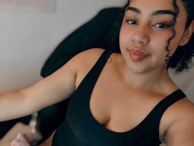 VickyCassel has light brown skin, curly black hair, and is wearing a black tank top, showing her small tits and slightly visible pussy, with a soft, confident smile.