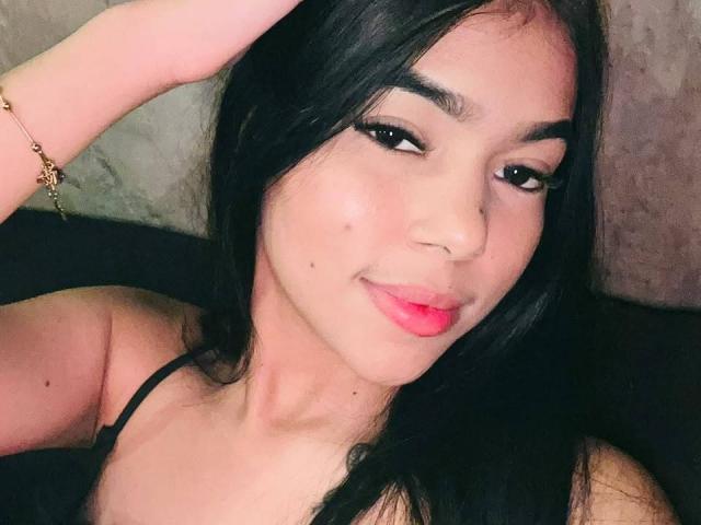 NataliaVelmonte with black hair, dark eyebrows, and pink lipstick, smiling slightly, wearing a black strap, hand on head, gold bracelet, light skin, cute face, small moles on cheek.