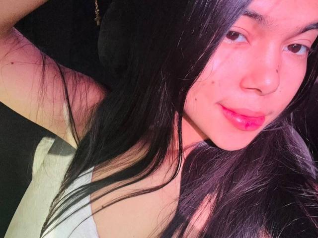 NataliaVelmonte has long black hair, red lips, and a few small acne spots on her pinkish skin. She's wearing a white top, looking at the camera with a slight smile.