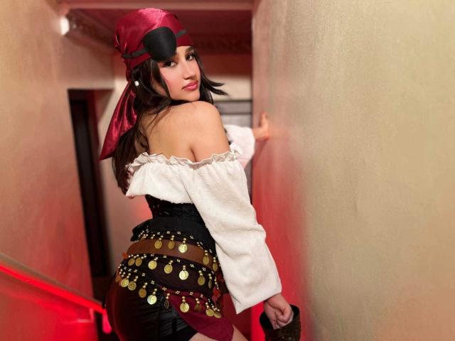 IrisBlair with dark hair, red eye mask, white off-shoulder top, black corset, and coin belt, leaning against a wall, looking back with a seductive smile, red lighting.