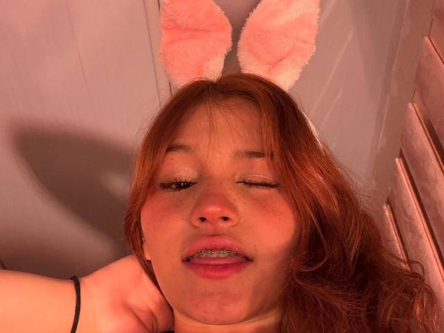 ElodieGardner with pink fuzzy bunny ears, winking, has red hair, freckles, and a slight open mouth showing teeth, wearing a black bracelet, in a warm light.
