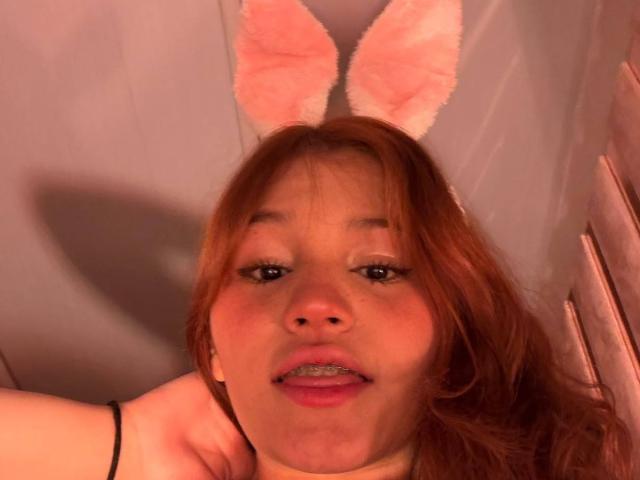 ElodieGardner with pink bunny ears, red hair, and a slightly open mouth, showing a small bit of her teeth, wearing a black bracelet, in a warm-lit selfie.