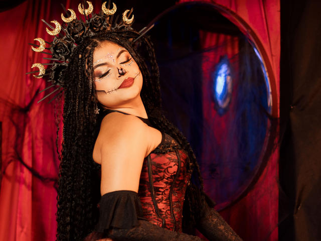 MelodyAlvarez with black braided hair, wearing a red lace corset, black gloves, and a spiked crown. Her eyes are closed, and she has dark makeup and red lipstick.