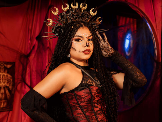 MelodyAlvarez with long black braids, red lace corset, black gloves, gold crown with crescent moons, and face paint, looking seductive, hand on head, dark red and black theme.