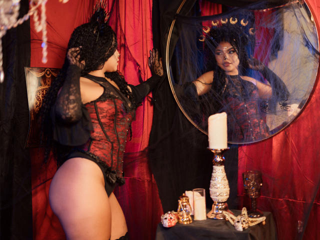 MelodyAlvarez in black lace headscarf and red-black corset, looking in mirror, showing her ass, wearing black panties, touching her face, with a candle and mask on table.