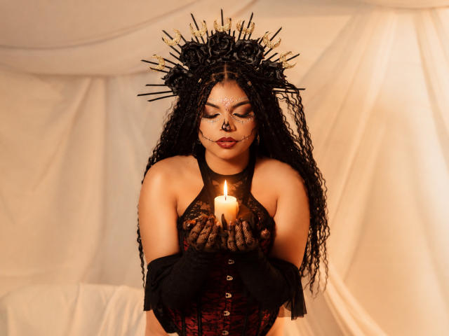 MelodyAlvarez with black braids, spiked headpiece, black lace top, holding candle, red corset, dark lipstick, and nose ring, gazing downward, warm light, soft fabric background.