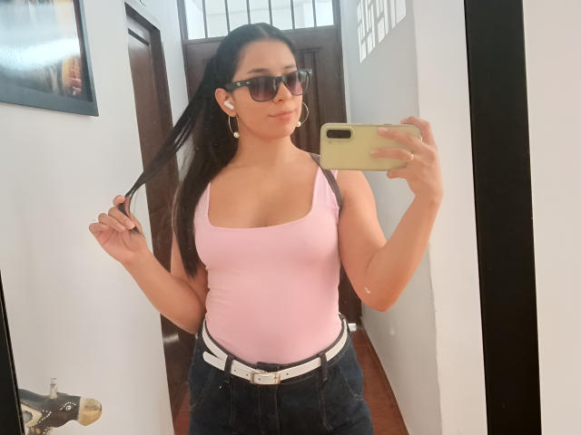 MelodyRoss with big boobs, pink tank top, dark jeans, white belt, black sunglasses, long black hair, holding hair, taking mirror selfie, iPhone in hand, white walls, wooden door.