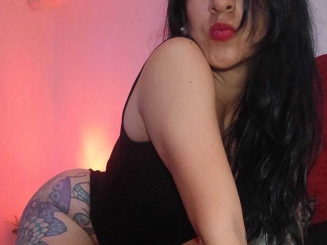 ZarayFeet, com cabelos pretos longos, batom vermelho, blusa de alça preta e tatuagens na coxa direita, inclinada para frente, fazendo beicinho, mostrando o bumbum. Luz rosa quente ao fundo..