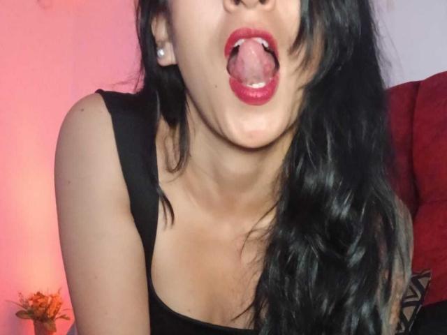 ZarayFeet with black hair, tongue out, red lipstick, black tank top, and earrings, showing her neck and upper chest, looking playful and sexy. Her tits are slightly visible.