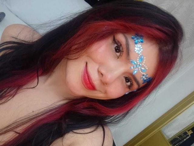 MaraCortes, con capelli rossi e neri, indossa gioielli floreali blu sul viso, sorride con rossetto rosso, ha gli occhi marroni e la pelle chiara, e nella foto mostra leggermente il seno..
