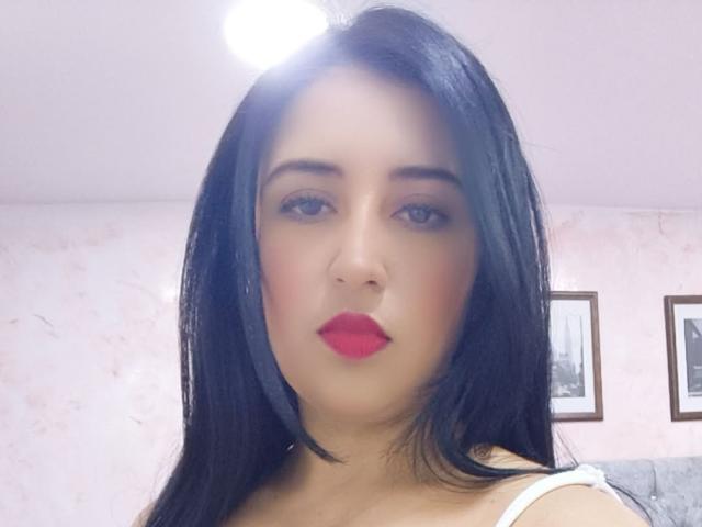 NicollJeans has straight black hair, red lipstick, and a neutral expression. She's wearing a white top with a visible strap. Her ass is not visible.