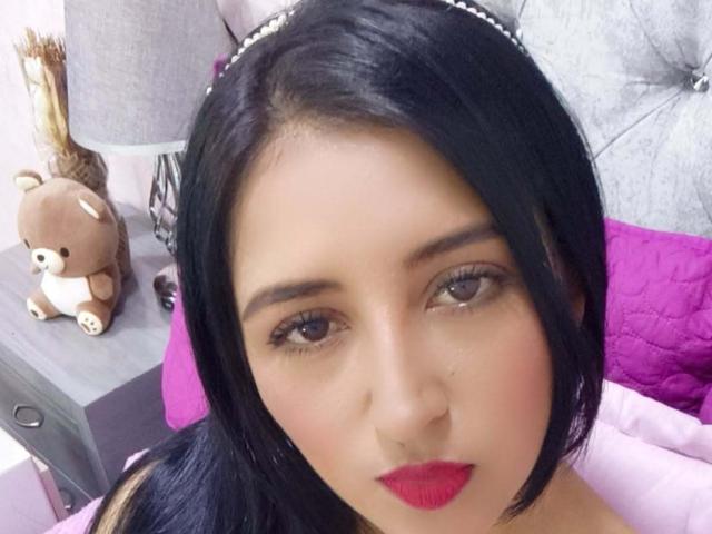 NicollJeans has black hair, red lipstick, and brown eyes. She's wearing a headband with a jeweled band. Her ass is not visible. A cute brown teddy bear is on a table.