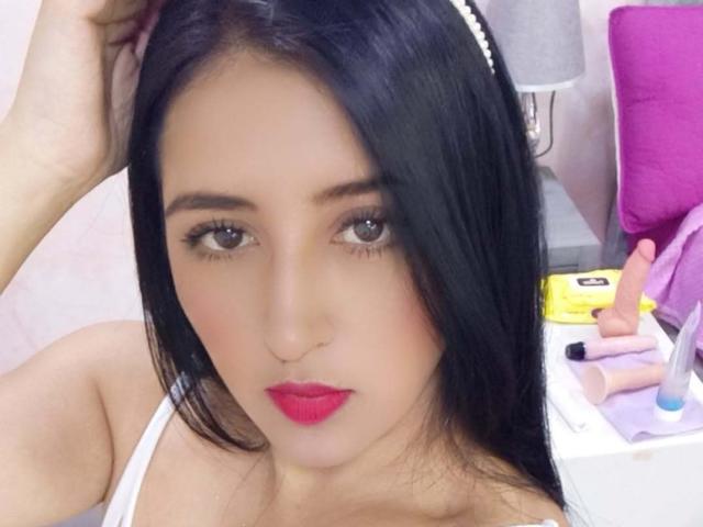 NicollJeans has black hair, red lipstick, and is looking at the camera with brown eyes. She's wearing a white headband and a white top, with a pink bag nearby.