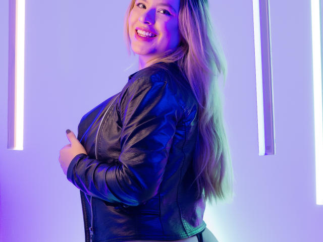 AshleySofia with long blonde hair, smiling, wearing a black leather jacket, hands on chest, purple and blue neon lights, showing her tits, standing sideways, looking over shoulder.