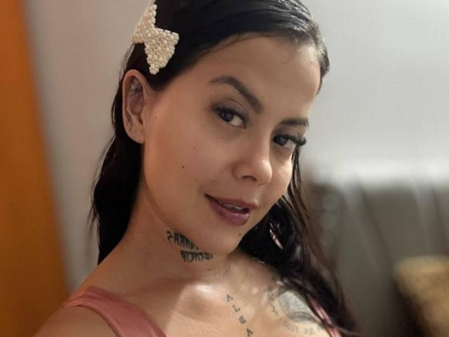 ElectraRoss has dark hair, a white jeweled hair clip, and a tattoo under her chin. She's looking at the camera with a slight smile, showing her tits.