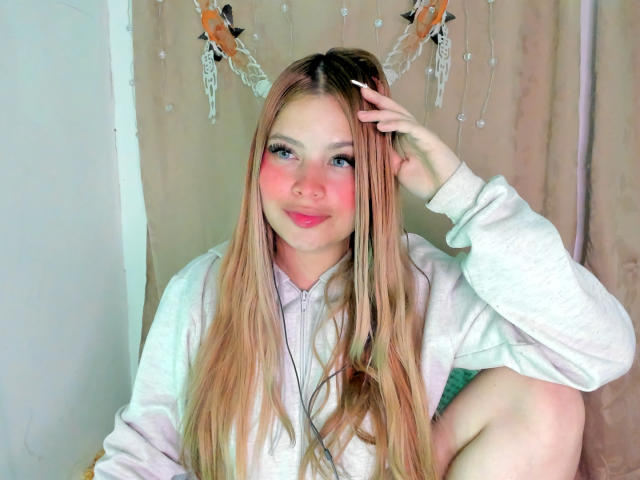 ShantalSummer has long blonde hair, pink cheeks, blue eyes, and is wearing a white hoodie with fake deer antlers. She's sitting with one knee up, hand in hair.
