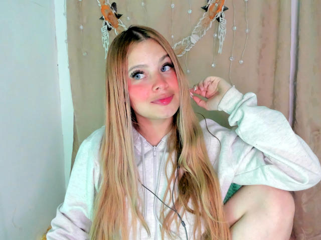 ShantalSummer has long blonde hair, pink blush on her cheeks, and is wearing a white hoodie. She's posing with one hand near her face, looking up with a cute smile.