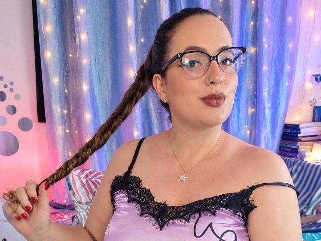 StacyMillerr with glasses, braided brown hair, holding a braid, wearing a purple lace-trimmed top, and a star necklace, has red lipstick and a cute smile.