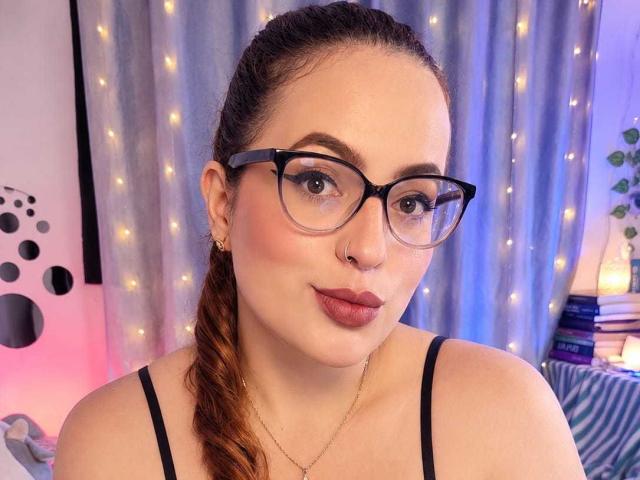 StacyMillerr with glasses, brown braid, nose ring, and red lipstick, wearing a black spaghetti strap top, has a cute face and small tits, smiling in a cozy room.