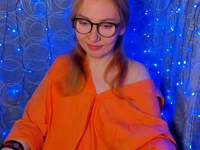 PaulinaSunny with glasses, orange off-shoulder top, and blonde pigtails, smiling subtly, blue-lit background with circular patterns and tiny lights. She's looking down, slightly blushing.