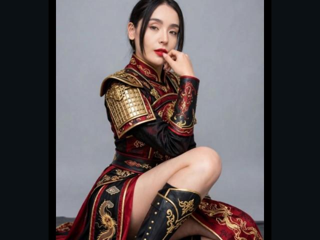 MindyLove, avec ses cheveux noirs, son rouge à lèvres rouge et son expression sérieuse, portant une armure rouge et or ornée, touchant ses lèvres avec son doigt, assise avec une jambe levée..