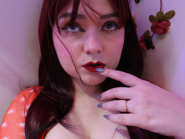 MaddyJones, avec du rouge à lèvres rouge, un vernis à ongles gris et une bague en or, touche ses lèvres rouges avec son doigt, a les cheveux roux, une peau pâle et un joli visage, et regarde l'appareil photo..