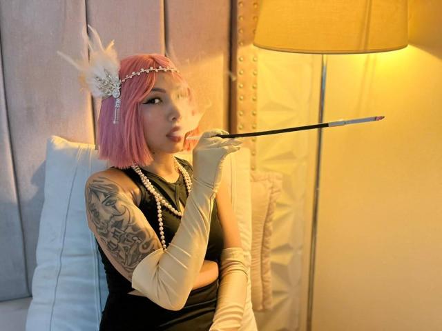 TracyLambo met een roze bob, een hoofdtooi met witte veren, een zwarte jurk, witte handschoenen, een parelketting, die een wandelstok vasthoudt, met haar tong uitgestoken, een getatoeëerde arm, sexy, onder een gele lamp, in een slaapkamer setting..