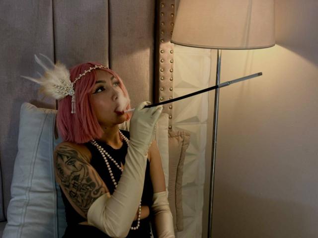 TracyLambo met een roze bob, een hoofdtooi met witte veren, een zwarte jurk, een parelketting, witte handschoenen, die een zwarte wandelstok vasthoudt, erop zuigt, een getatoeëerde arm, een lamp op de achtergrond, kijkt omhoog..