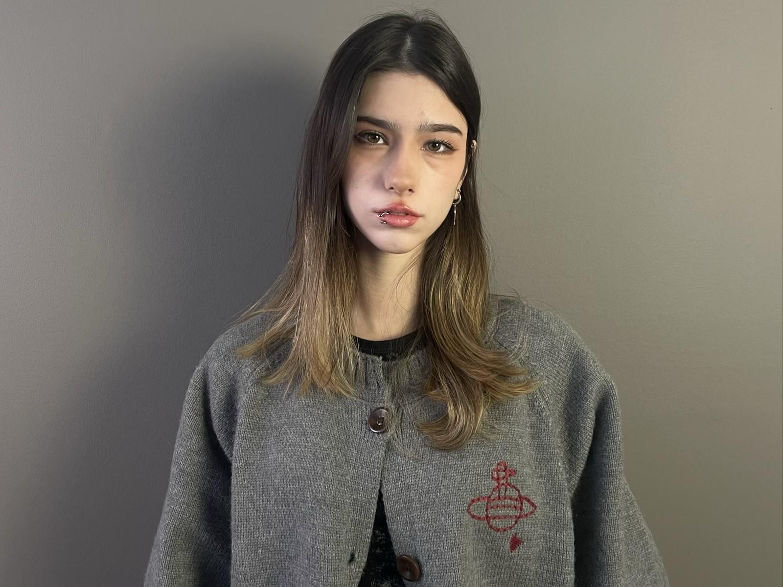 Dyan Herriott, com cabelos castanhos longos, um casaco cinza abotoado com bordado vermelho e um piercing no nariz, olhando para a câmera. Ela tem um pequeno piercing no lábio e uma expressão neutra..