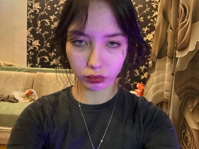 JovanLockett, con i capelli neri e disordinati, una luce viola sul viso, una piccola lacrima sotto l'occhio, una maglietta nera, una collana, labbra piene e un'espressione stanca, seduto in una stanza disordinata..
