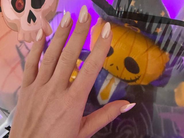 AlicePrings' hand with long, nude-painted nails, holding a yellow plush toy with black eyes and a smile, next to a pink skull toy with black eyes.