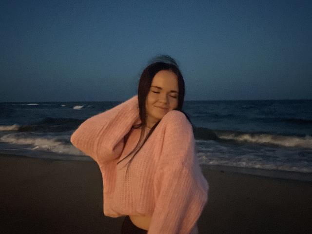 RoxyVesper with long brown hair, closed eyes, smiling, wearing a pink knit sweater, hands in hair, standing on a beach at dusk, ocean and waves in background.