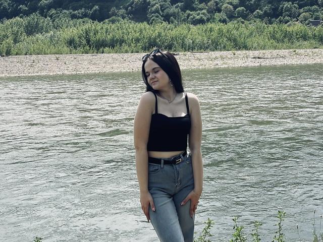 RoxyVesper with black crop top, small tits, blue jeans, and belt, standing by a river, smiling, arms relaxed, long black hair, sunglasses on head, green foliage in background.