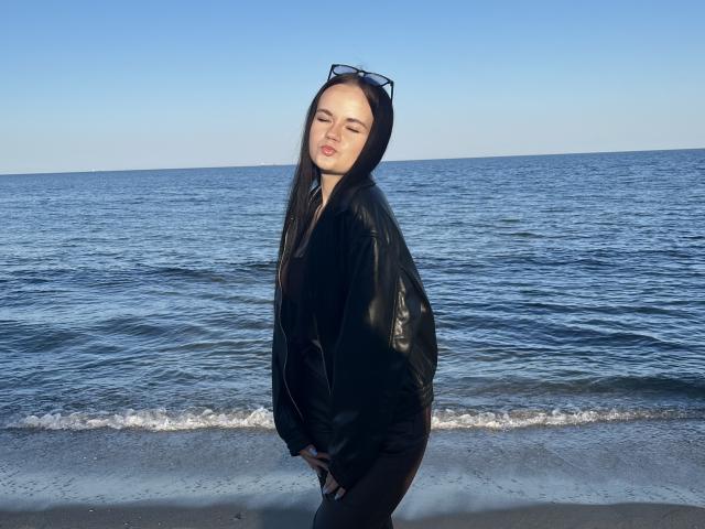 RoxyVesper with long black hair, eyes closed, wearing a black leather hood and black pants, standing on a beach with blue ocean in the background, sunglasses on head.