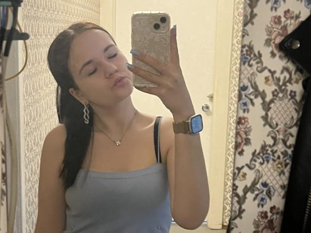 RoxyVesper, with pouty lips, takes a mirror selfie, wearing a grey tank top, black bra straps, silver earrings, and a gold watch, holding an iPhone with a floral case.