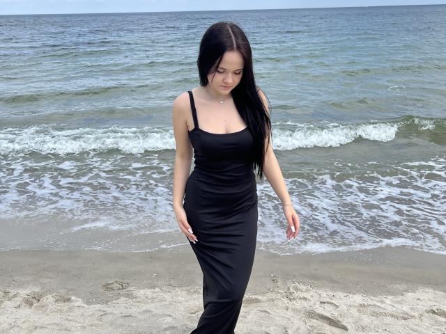 RoxyVesper with long black hair, wearing a tight black dress, standing on a sandy beach, looking down, her small tits and slender ass visible, ocean in background.